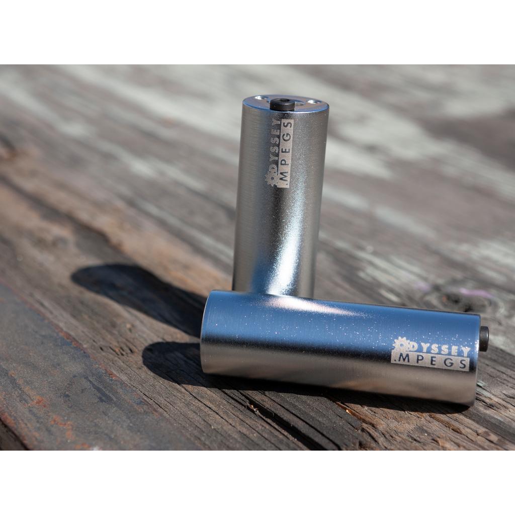 Two Odyssey MPEG Pegs Pair, both metallic, rest on a weathered wooden surface—one peg stands upright while the other lies horizontally.