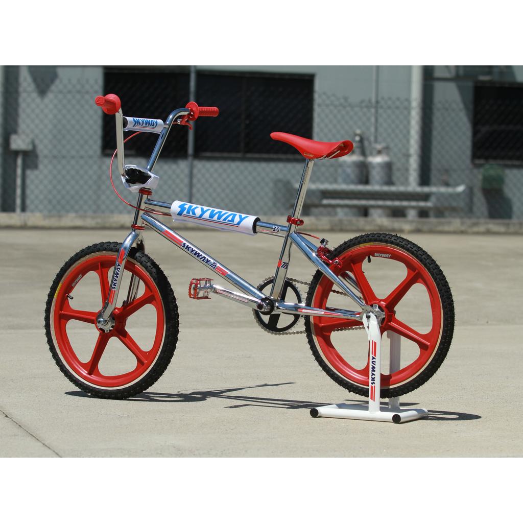 A silver BMX bike with red wheels and seat is showcased on a Skyway Stolz Stand outdoors by a chain-link fence.