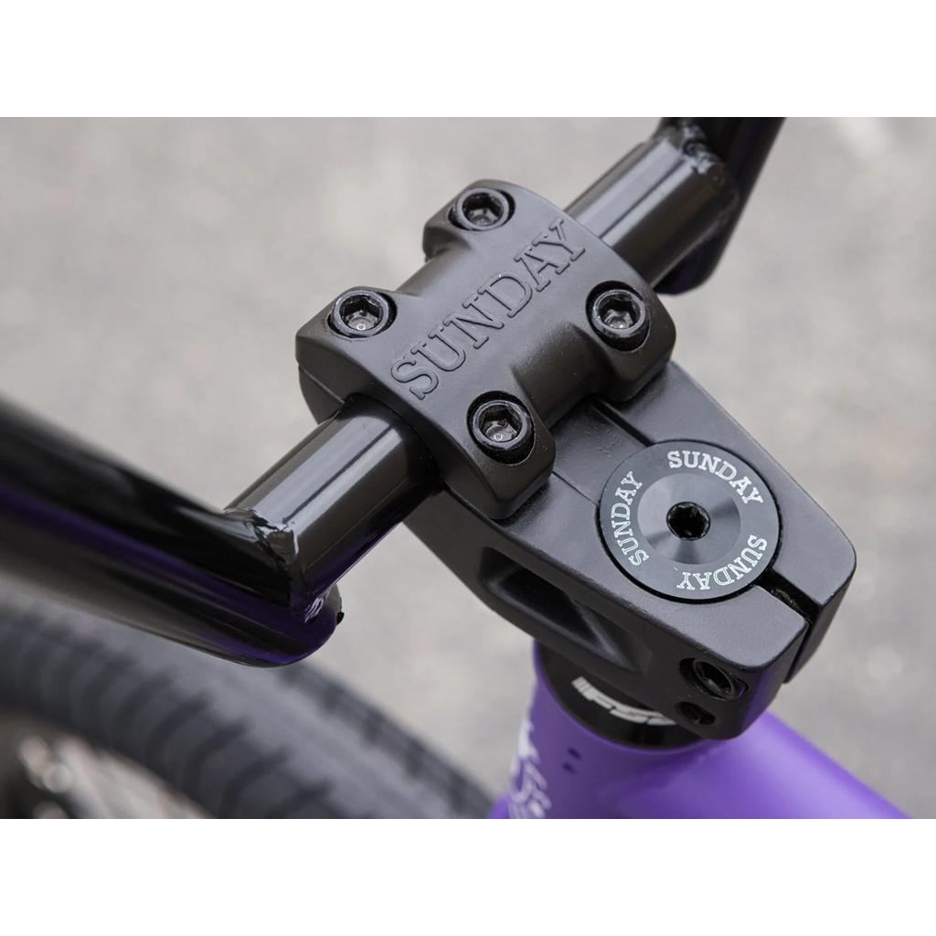 Close-up of a black handlebar stem with "SUNDAY" engraved on it, attached to a purple Sunday Forecaster 20 Inch Bike featuring Odyssey parts.