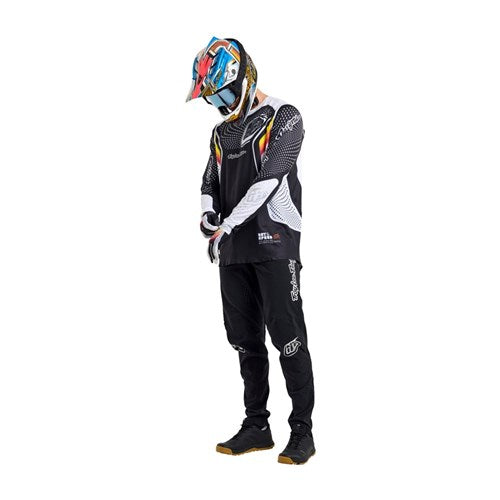 A person in a full motocross outfit with a colorful helmet, Precision Fit black and white jersey, TLD 26.1 Sprint Ultra Pant / Mono Black, gloves, and shoes stands looking downward.