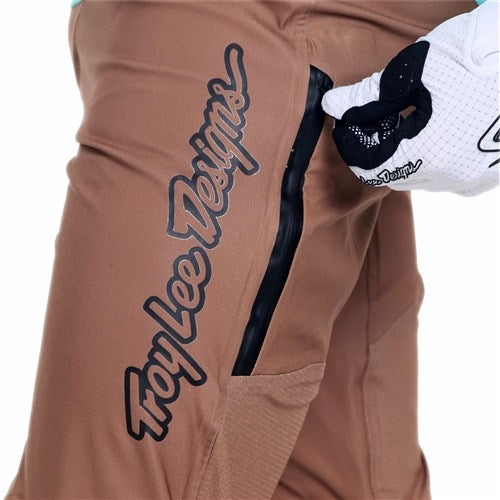 A gloved hand unzips a side pocket on the brown TLD 26.1 Sprint Ultra Pant / Mono Sienna, featuring the "Troy Lee Designs" logo in black and showcasing the lightweight design of premium racing gear.