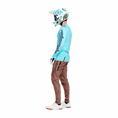 A person in Troy Lee racing gear, including a teal helmet and jersey, TLD 26.1 Sprint Ultra Pant in Mono Sienna, white gloves, and shoes stands sideways against a white background.