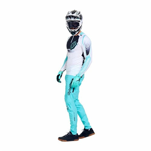 Someone stands against a white background wearing the TLD 26.1 Sprint Ultra Pant in Mono Real Teal with gloves, black shoes, and a full-face helmet; the outfit features Bluesign-approved fabric.