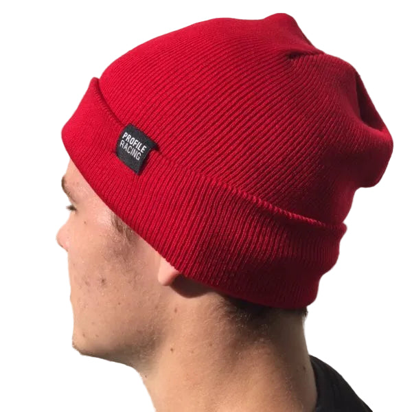 A person stands in sunlight against a light brick wall, wearing the Profile Woven Label Beanie—a bright red knit beanie with a "Profile Racing" woven label.
