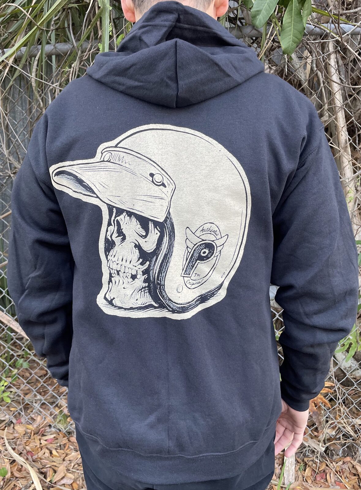 Wearing the Profile From the Dungeon Zip Hoodie, featuring a large skull in a motorcycle helmet by Adi Gilbert on the back, this person stands before a chain-link fence and foliage.