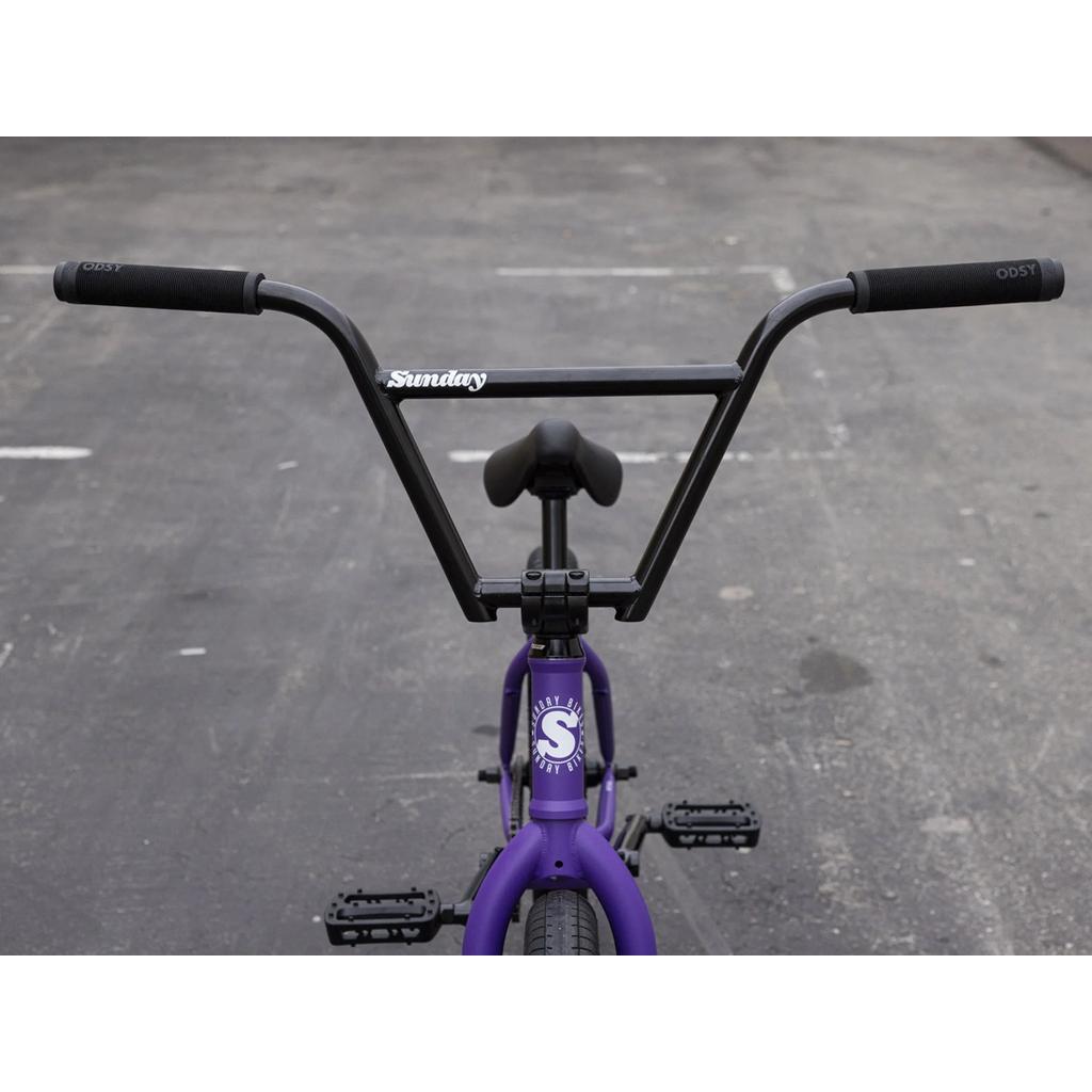 A close-up of the handlebars and front chromoly frame of the purple Sunday Forecaster 20 Inch Bike with Odyssey parts, set against an empty asphalt background.