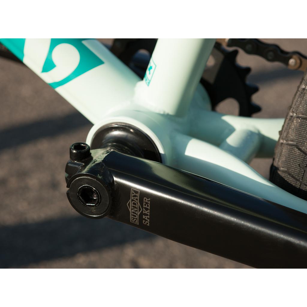 Close-up of a Sunday Saker crank arm on a light blue 100% chromoly frame from the Sunday EX Julian Arteaga 20 Inch Bike, with rear wheel and chain in the background.