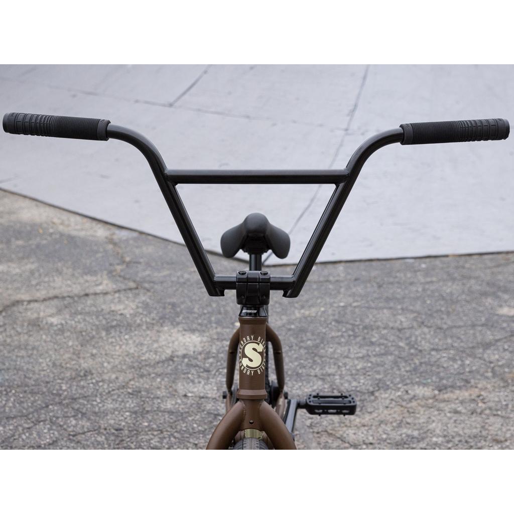 Close-up of the handlebars, seat, and front of a brown Sunday EX Julian Arteaga 20 Inch Bike, made from 100% chromoly, shown outdoors on concrete.