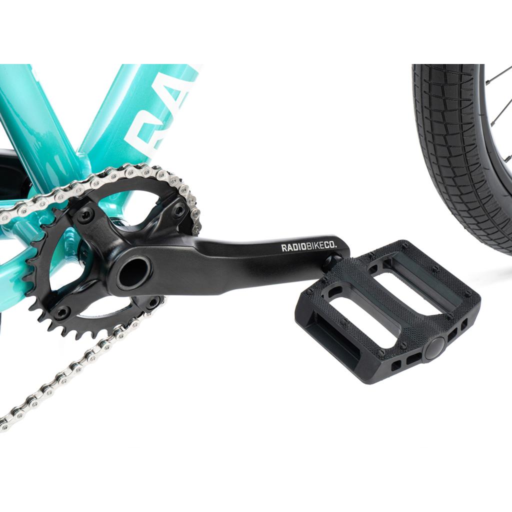 Close-up of a black crank arm and flat pedal attached to the chainring and turquoise 6061-T6 alloy frame of the Radio Fiend 26 Inch Bike, with part of the rear wheel visible.