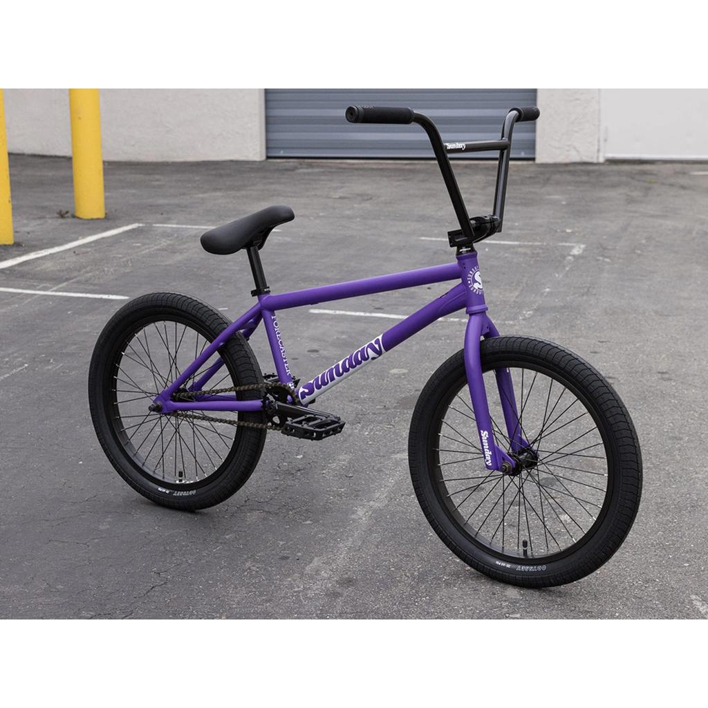 A purple Sunday Forecaster 20 Inch Bike with black handlebars and tires is parked on asphalt near yellow poles and a closed garage door.