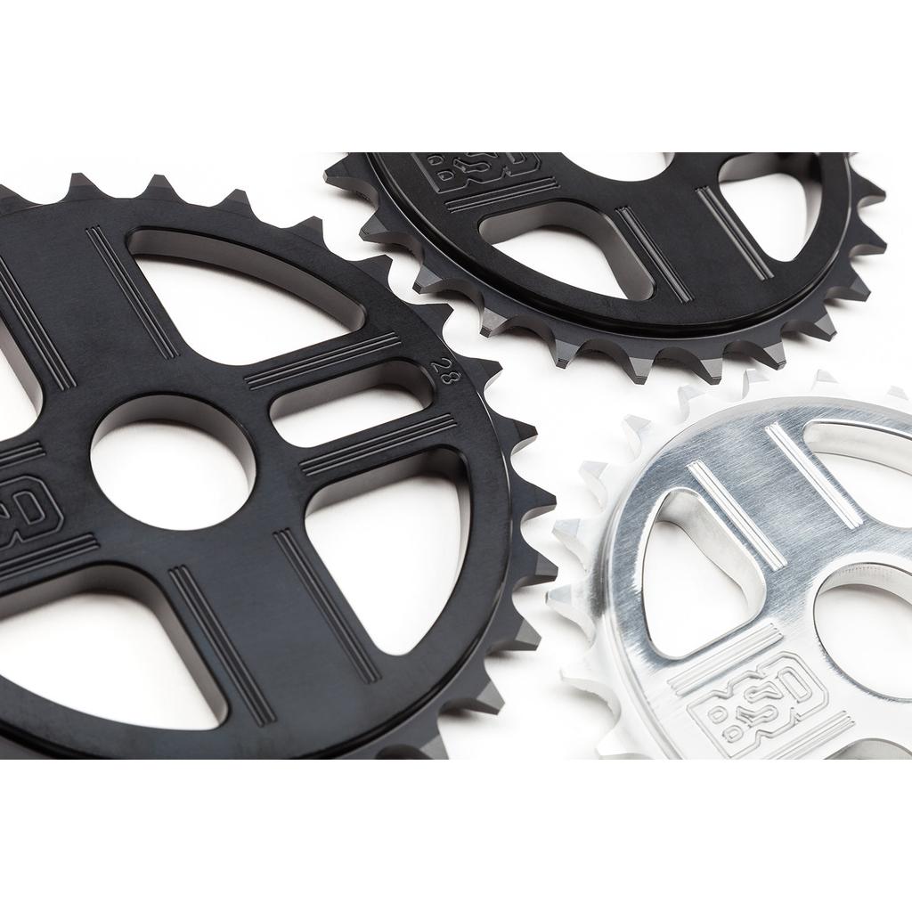 Three bicycle chainrings, including a BSD TBT Sprocket made of durable 7075 aluminium, are displayed on a white surface—two are black and one is silver.
