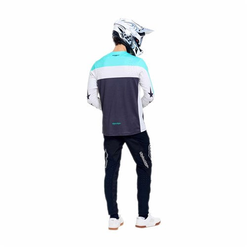 A person stands facing away, wearing a TLD 26.1 Sprint Pro Jersey / Yo Moto Carbon in teal, white, and gray with Race Fit design and black motocross pants.