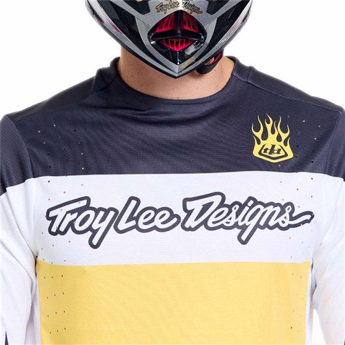 A person wears the TLD 26.1 Sprint Pro Jersey / Yo Moto Butter in black, white, and yellow with a helmet; the jersey features a flame logo on the upper left chest and moisture-wicking fabric for comfort.