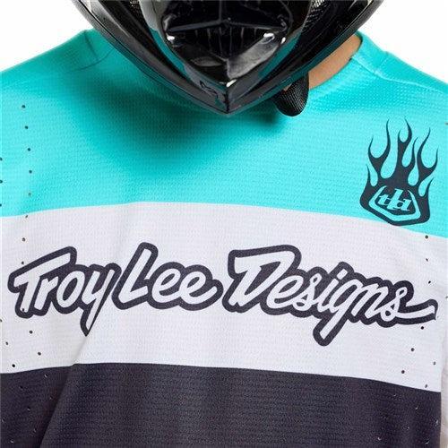 Close-up of a person in a black helmet wearing the TLD 26.1 Sprint Pro Jersey (Youth) / Yo Moto Carbon, shown in turquoise, white, and black with a flame logo on the chest and featuring a sleek Race Fit.