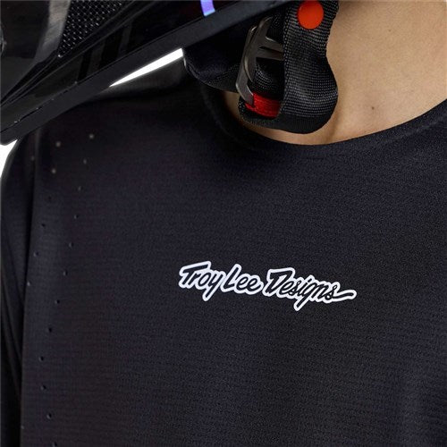 Close-up of a person wearing a TLD 26.1 Sprint Pro Jersey (Youth) / Mono Black and a black helmet with a red chin strap.