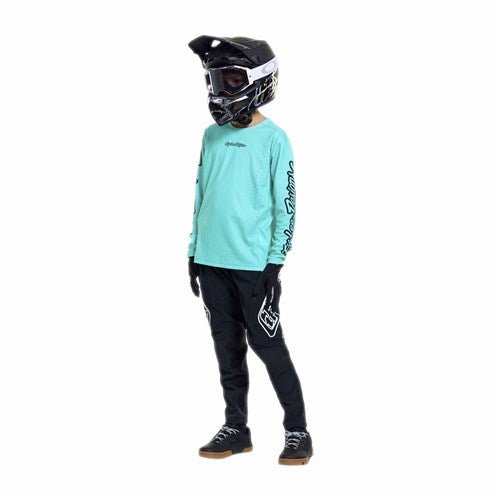 A person wearing a TLD 26.1 Sprint Pro Jersey (Youth) / Mono Real Teal, black pants, gloves, and a black motocross helmet stands against a plain white background.