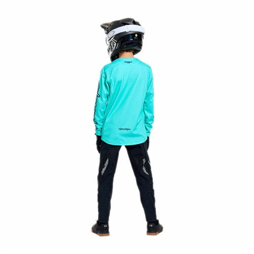 A child in a TLD 26.1 Sprint Pro Jersey (Youth) Mono Real Teal, black pants, gloves, and a black-and-white helmet stands facing away from the camera against a white background.