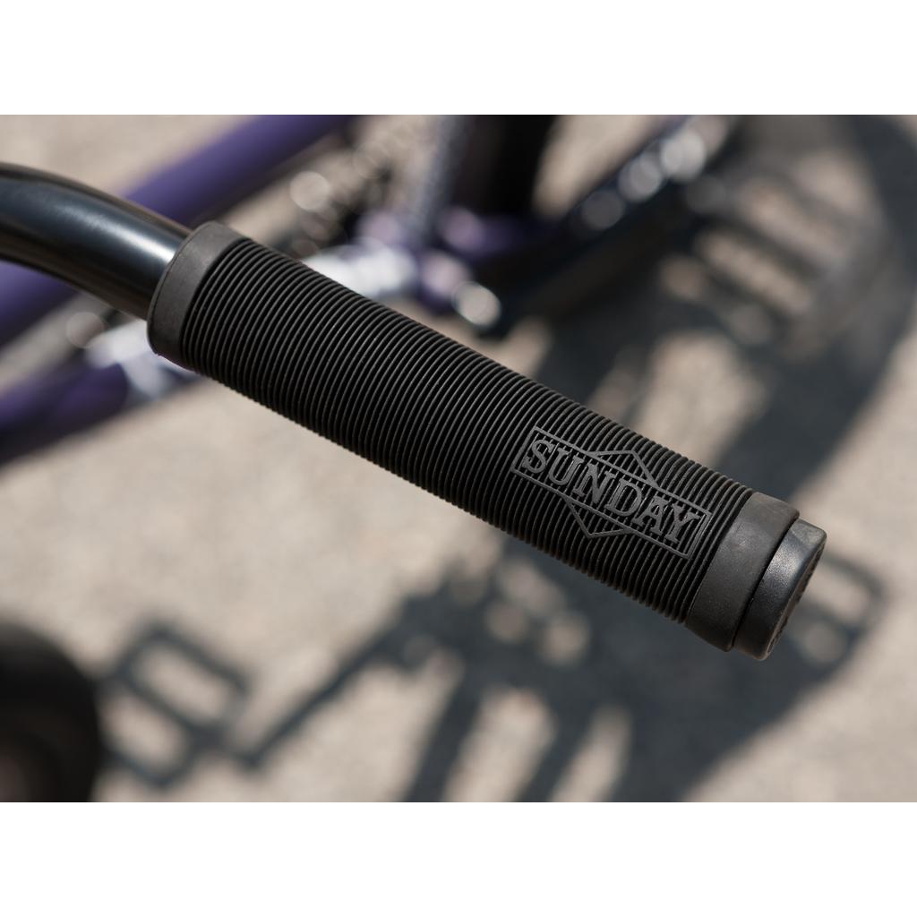 Close-up of a black handlebar grip embossed with "SUNDAY," mounted on a Sunday Forecaster Maca Grasset 20 Inch Bike outdoors.