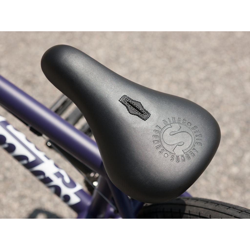 Close-up of a black bicycle seat with the "Sunday Bikes" logo and signature embossed, mounted on a purple Sunday Forecaster Maca Grasset 20 Inch Bike frame.
