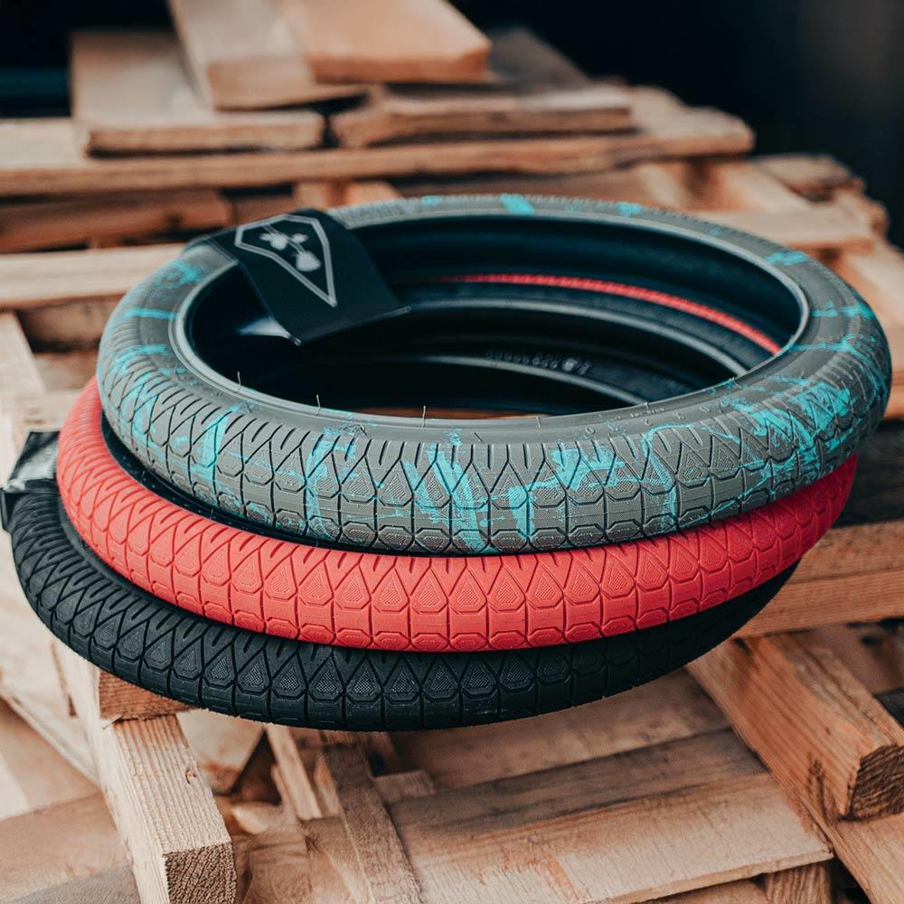 Three BMX street tyres, including the turquoise and black Matt Ray signature tyre, a red Subrosa Designer Tyre, and a black option, are stacked neatly on wooden pallets.