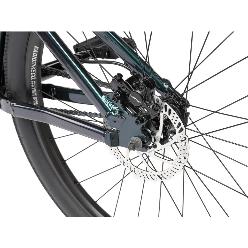 Close-up of the Radio Griffin 26 Inch Bike’s rear wheel, featuring Tektro disc brakes with rotor and caliper, plus spokes and a section of the Maxxis DTH tire.