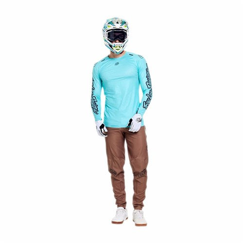 Person wearing a TLD 26.1 Sprint Ultra Jersey in Mono Real Teal made with moisture-wicking Ultra Knit fabric, brown pants, white gloves and shoes, and a motocross helmet, standing against a plain white background.