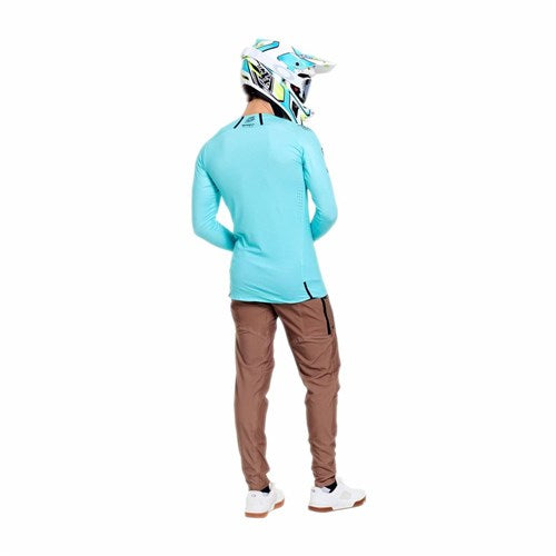 A person wearing a TLD 26.1 Sprint Ultra Jersey Mono Real Teal, brown pants, white shoes, and a white and blue helmet stands facing away from the camera.