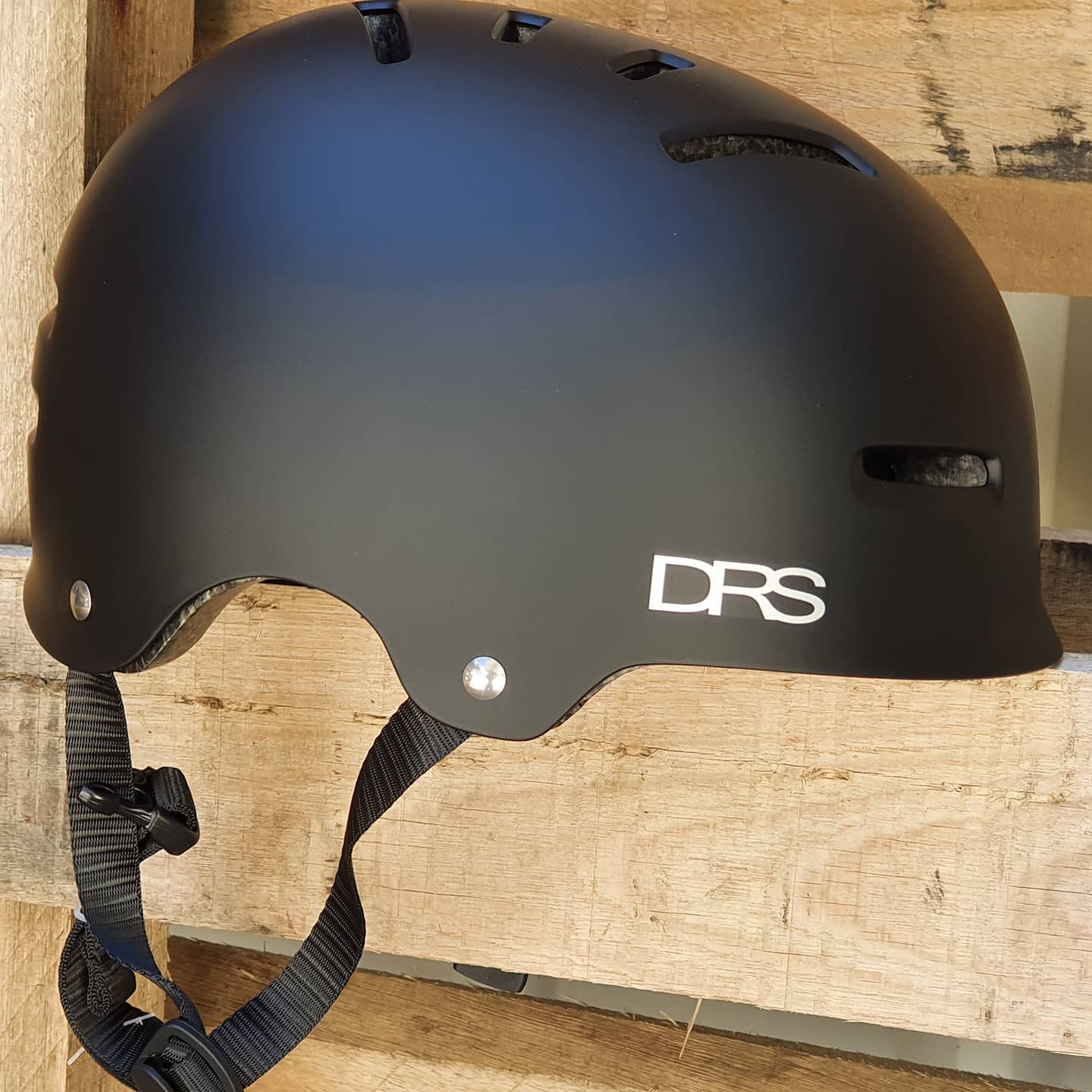 The DRS Helmet Flat Black, featuring a chin strap, rests on a wooden pallet. It meets Australian Standards for safety and style.
