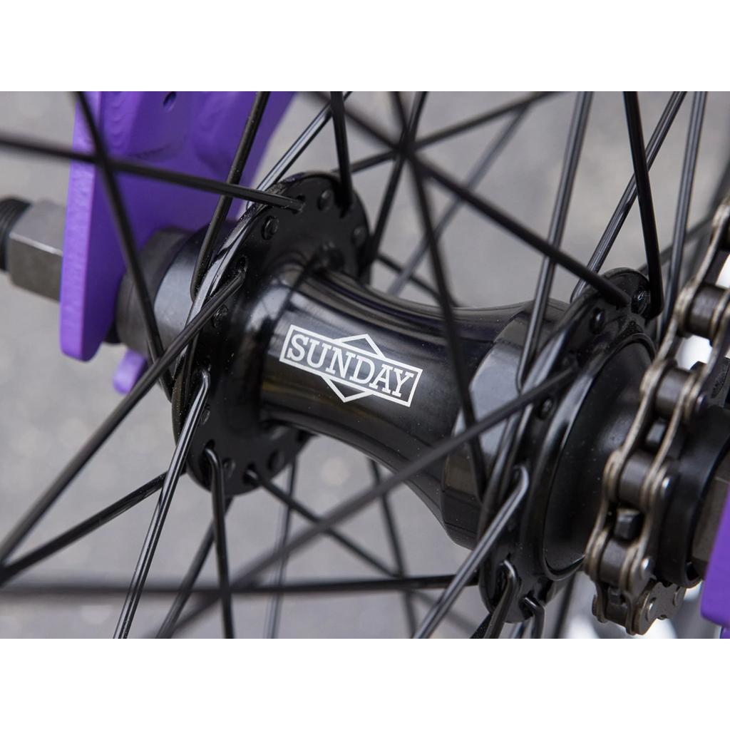 Close-up of a black hub with "SUNDAY" on it, black spokes, and part of the purple chromoly frame of the Sunday Forecaster 20 Inch Bike, highlighting high-quality BMX components.