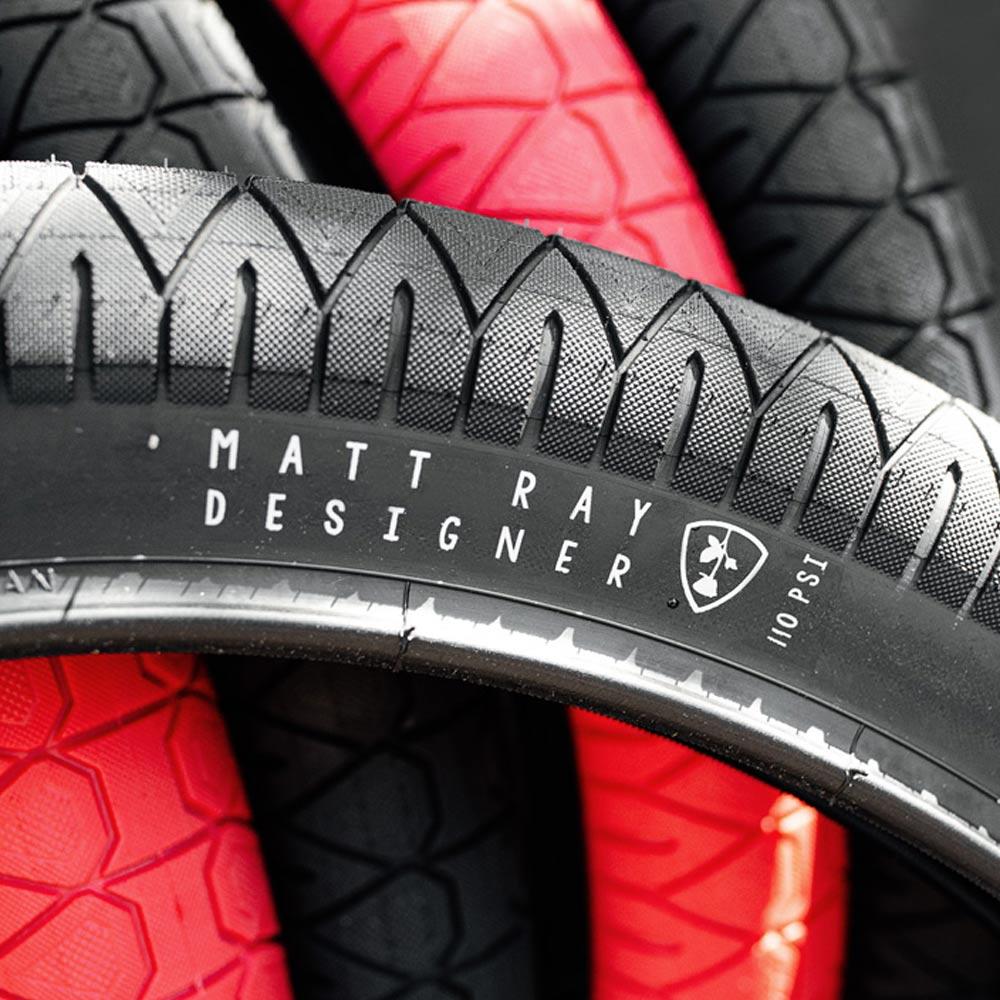 Close-up of black and red Subrosa Designer Tyres stacked, showing "Matt Ray Designer," "110 PSI," and branding on the black Matt Ray signature tyre's sidewall.