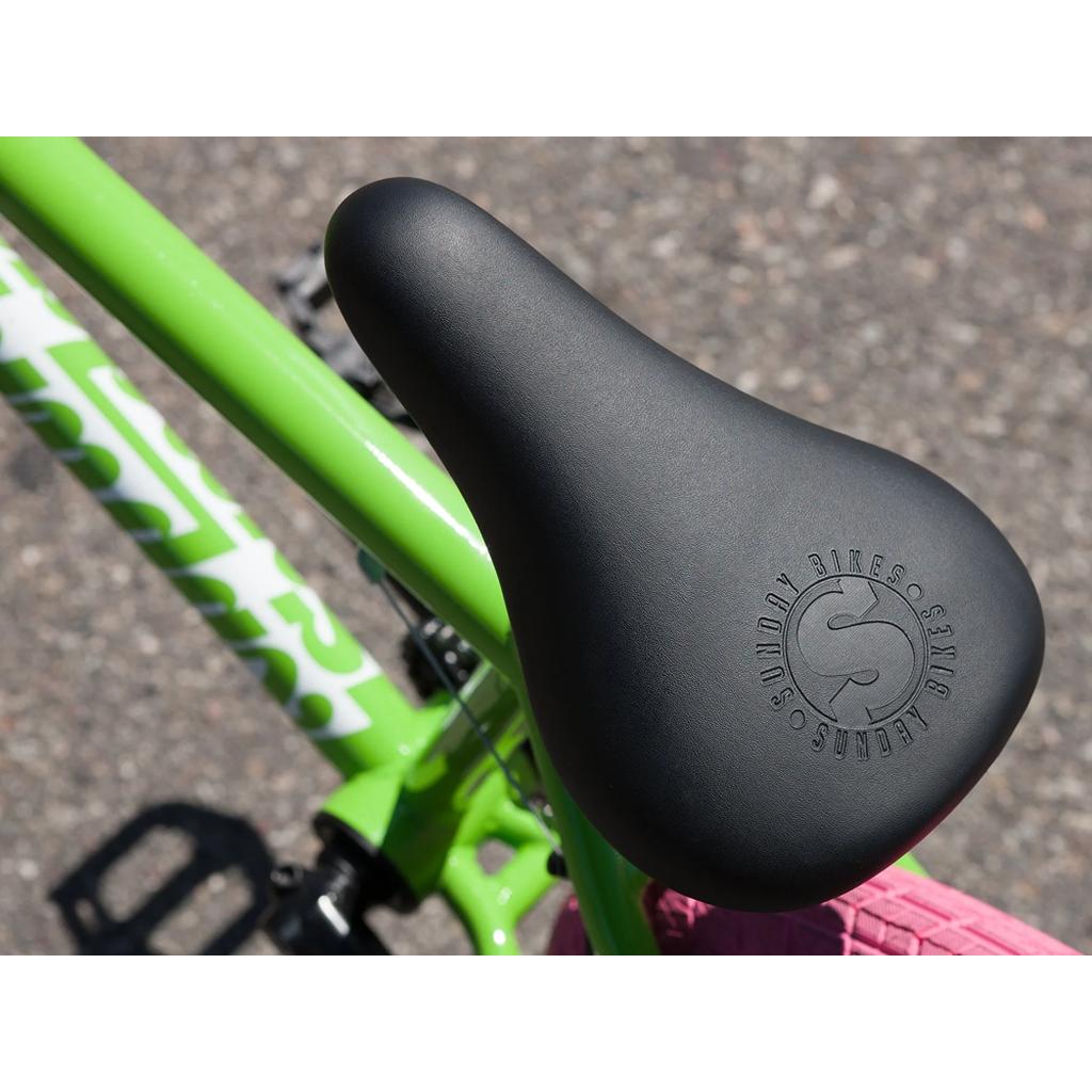 Close-up of the Sunday Blueprint 20 Inch Bike (2023): black seat with "Sunday Bikes" logo, mounted on green frame with pink handlebar—offers pro-level geometry ideal for riders wanting more than a beginner’s bike.