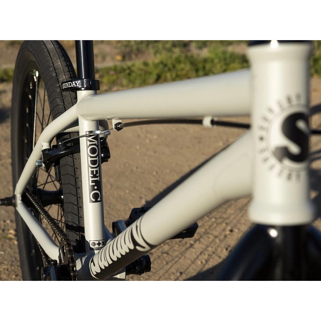 Close-up of the Sunday Model C 24 Inch Bike in light gray and black, featuring a Chromoly frame, quality Odyssey parts, and "MODEL C" and "SUNDAY" logos, photographed outdoors on a dirt path.