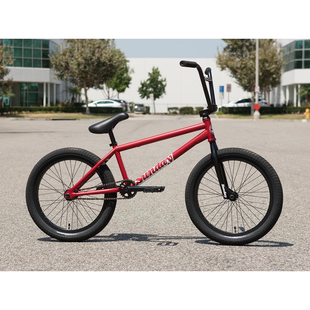 A Sunday Forecaster 20 Inch Bike in red, featuring black handlebars, seat, and tires, is parked on a paved area with office buildings and trees in the background.