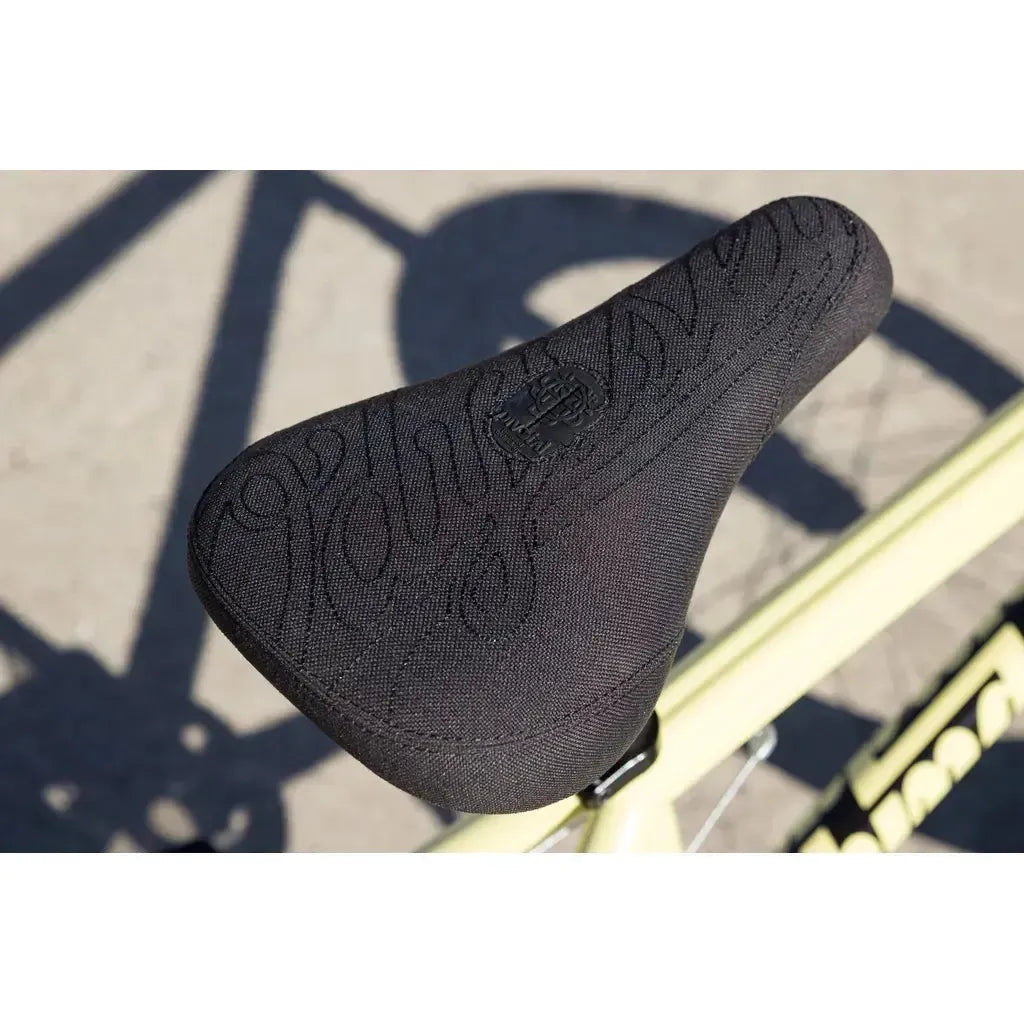 Close-up of a black, textured bicycle seat on the light yellow frame of the 2025 Sunday Soundwave Special Bike, equipped with quality Odyssey parts and set against a blurred pavement background.
