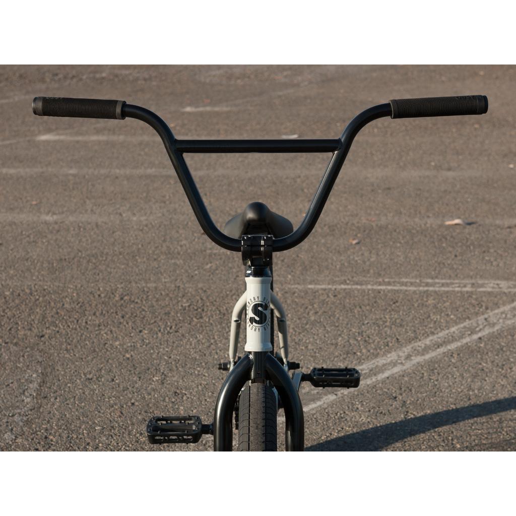 A front view of the black Sunday EX Erik Elstran 20 Inch Bike with wide handlebars, black pedals, a white logo on its 100% chromoly frame, and Odyssey Springfield Brakes, shown on an empty asphalt surface.