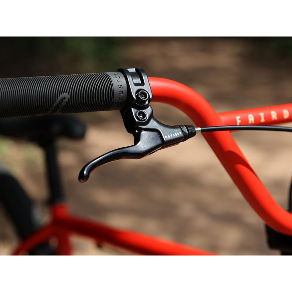 Close-up of a black brake lever on a red handlebar, with "FAIRD" partly visible on the Fairdale Taj 27.5 Inch Bike (2023) frame—a sleek single speed ideal for BMX-style commuting.