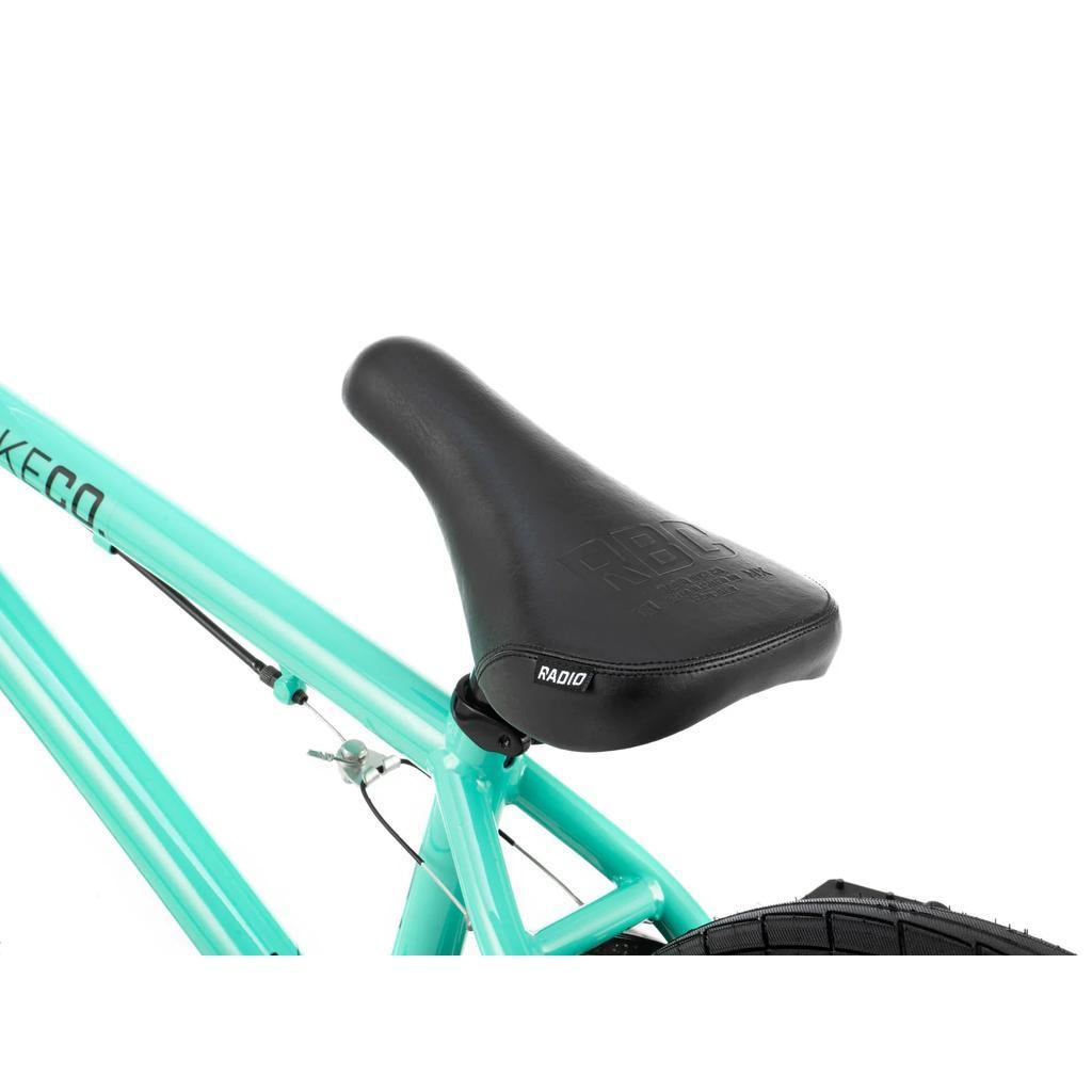 Close-up of the black "RADIO" seat on the Radio Saiko 18 Inch Bike in teal, featuring part of the rear wheel and brake cable—ideal for young BMX fans.