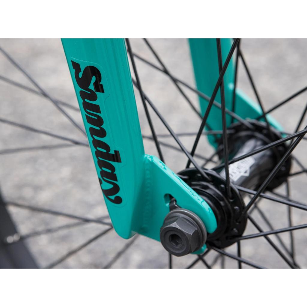 Close-up of the Sunday EX Erik Elstran 20 Inch Bike’s turquoise fork with black "Sunday" branding, black spokes, and axle on blurred pavement. Designed for strength, this fork complements Odyssey parts on a rugged chromoly frame.