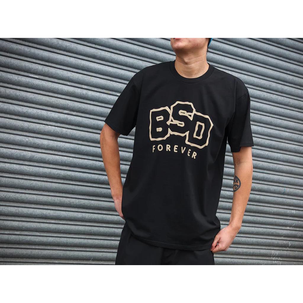A person stands before a corrugated metal wall wearing the BSD Wobbler T-Shirt by Ninety One, with "BSD FOREVER" text, and has a tattoo on their right forearm.