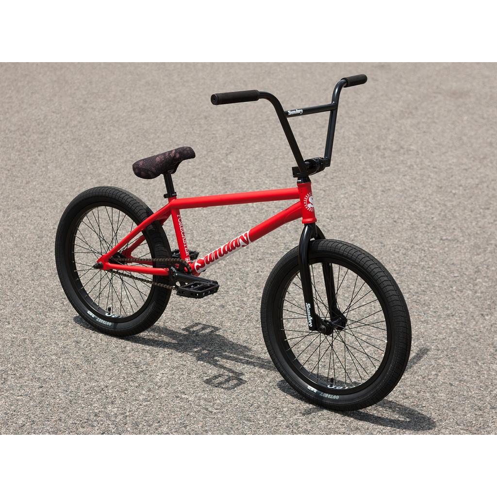 A Sunday Forecaster 20 Inch Bike (2025) / Brett Silva with a red 100% chromoly frame, black handlebars, wheels, and pedals stands upright on asphalt.