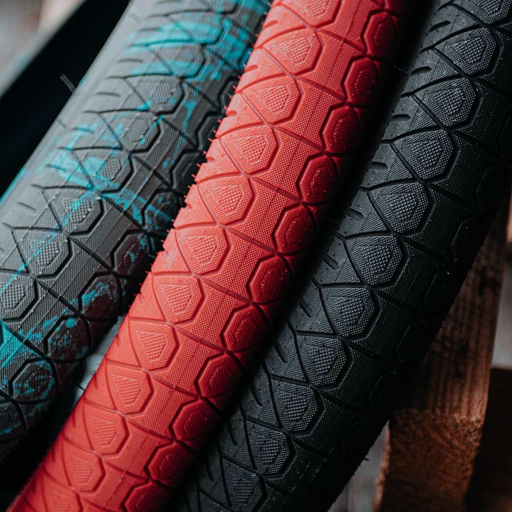 Three Subrosa Designer Tyres in black, red, and black with blue splatters, each with a hexagonal tread, are lined up closely—highlighting this BMX street tyre.