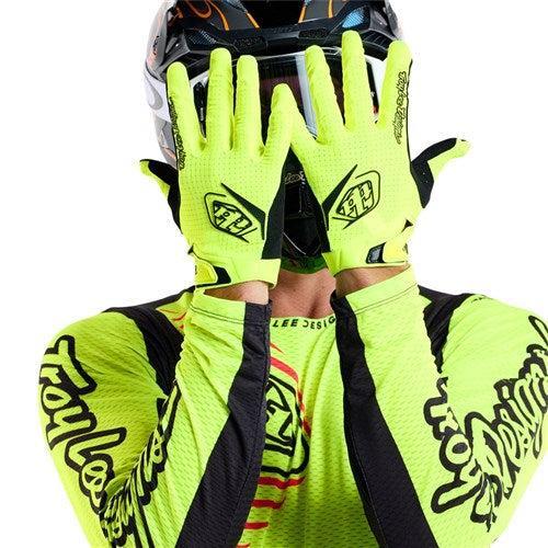 A person in a neon yellow and black biking outfit and helmet hides their face with TLD 26.1 Air Gloves (Youth) in Flo Yellow, featuring touchscreen compatibility, ergonomic fit, and Troy Lee Designs logos.