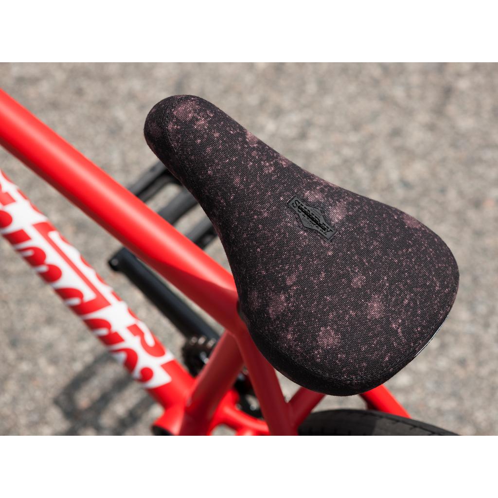 Close-up of a black and pink speckled bicycle seat on a Sunday Forecaster 20 Inch Bike (2025) / Brett Silva with a 100% chromoly frame, set against a blurred pavement background.
