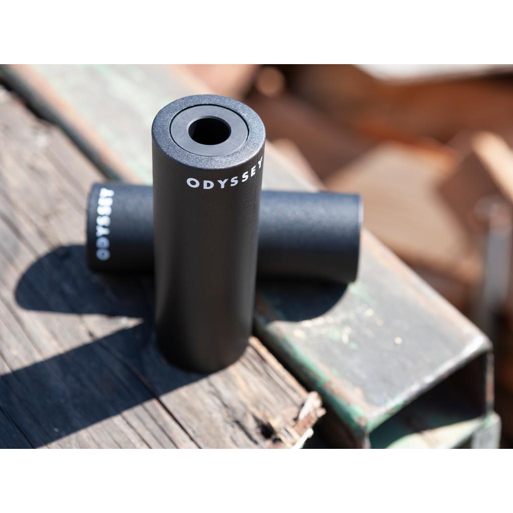A single black cylindrical Odyssey Joystick PC Peg rests on a weathered wooden surface outdoors, featuring a durable lightweight aluminum core.