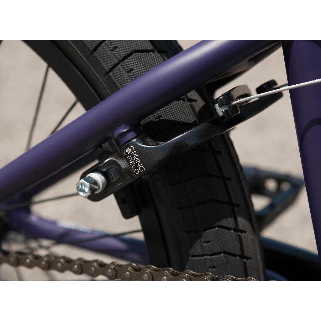Close-up of a bicycle's rear wheel showing a Springfield brake caliper, tire tread, chain, and part of the purple Sunday Forecaster Maca Grasset 20 Inch Bike frame.