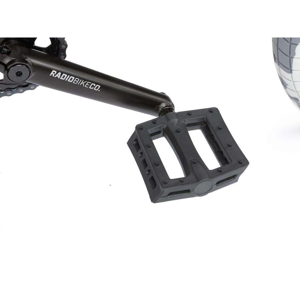 Close-up of a black pedal attached to a crank arm labeled "RADIOBIKECO," with part of the wheel from the Radio Darko 20 Inch Bike visible on the right.