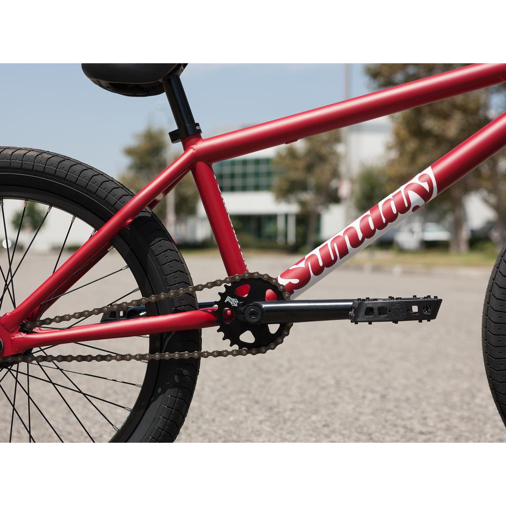 Close-up of the Sunday Forecaster 20 Inch Bike’s red chromoly frame with "Sunday" branding, Odyssey parts, chain, pedals, and rear wheel visible; blurred buildings and trees in the background.