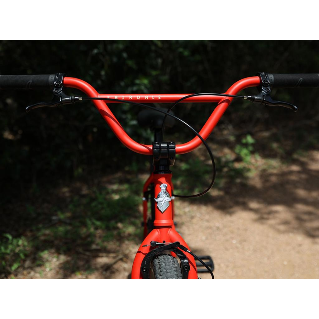 Close-up of the Fairdale Taj 27.5 Inch Bike (2023) handlebar and front wheel on a dirt path, surrounded by trees and foliage.