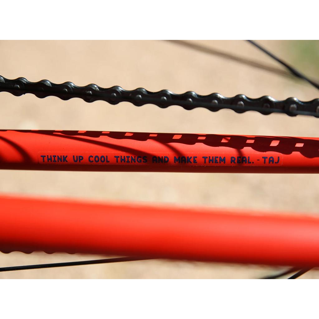 Close-up of a red Fairdale Taj 27.5 Inch Bike (2023) frame featuring the text “THINK UP COOL THINGS AND MAKE THEM REAL. - TAJ” on the tube, with a bicycle chain above—ideal inspiration for BMX commuters.