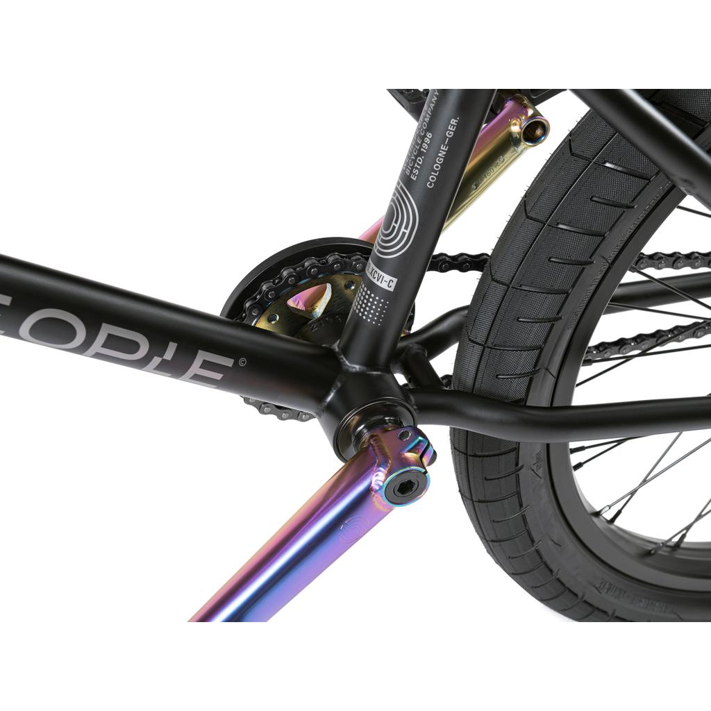 Close-up of the Wethepeople Reason Shift 20 Inch Bike's rear wheel, chain, and crank area, showing a black full chromoly frame, metallic rainbow crank arms, textured tire, and a hybrid freecoaster cassette setup.